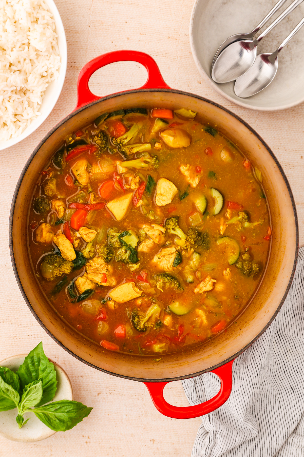 A red pot filled with chicken and vegetable curry sits on a beige surface, surrounded by a bowl of rice, a plate with spoons, fresh basil, and a striped napkin.