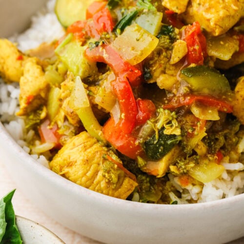 Bowl of white rice topped with colorful stir-fry made of chicken, red peppers, zucchini, onions, and broccoli. Fresh basil leaves are visible on a plate nearby.