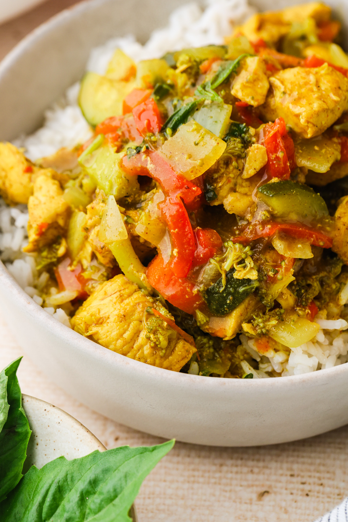 Bowl of white rice topped with colorful stir-fry made of chicken, red peppers, zucchini, onions, and broccoli. Fresh basil leaves are visible on a plate nearby.