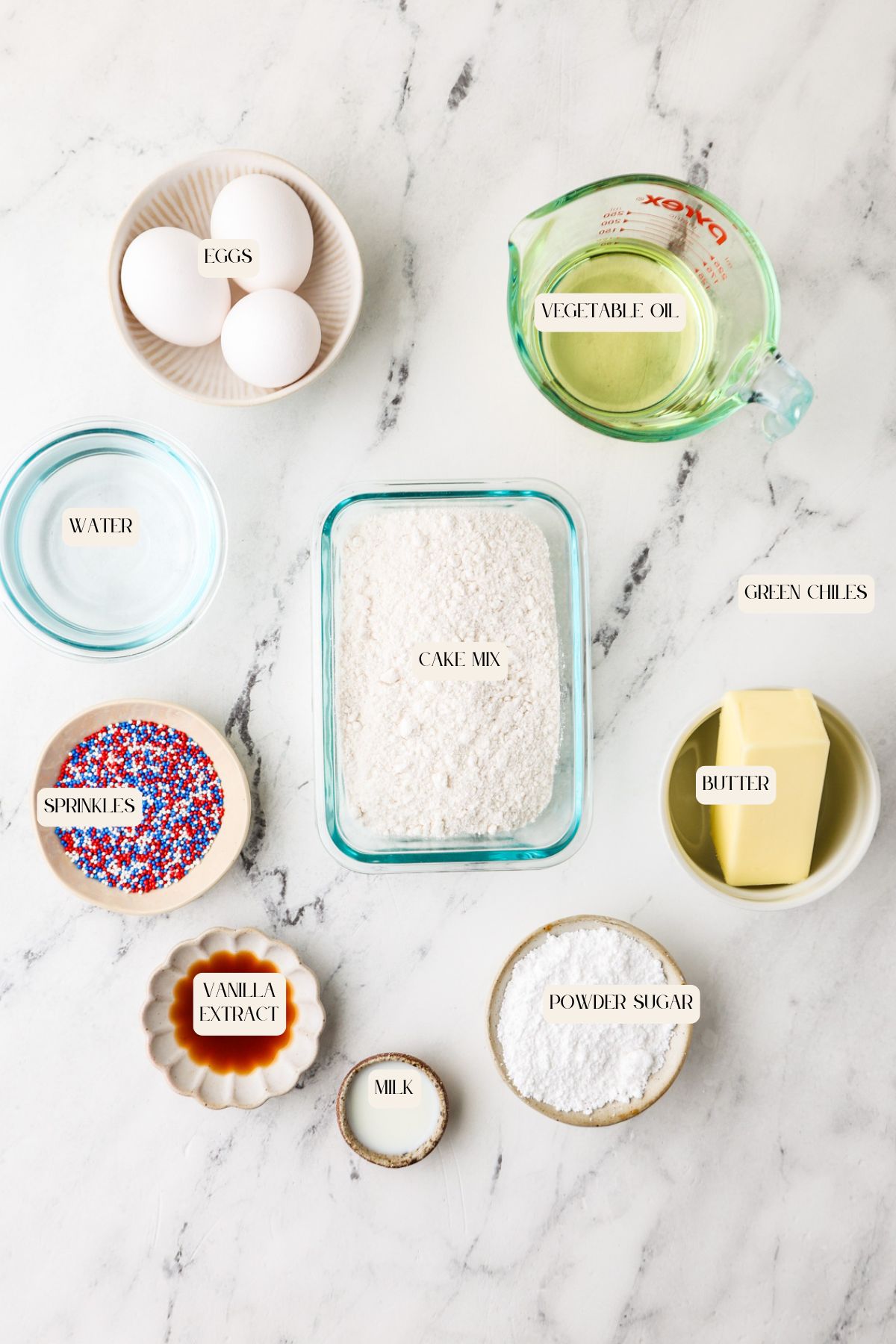 A marble countertop displays eggs, water, sprinkles, vanilla extract, cake mix, powdered sugar, milk, green chiles, butter, and vegetable oil in bowls and cups, each labeled with its ingredient name.
