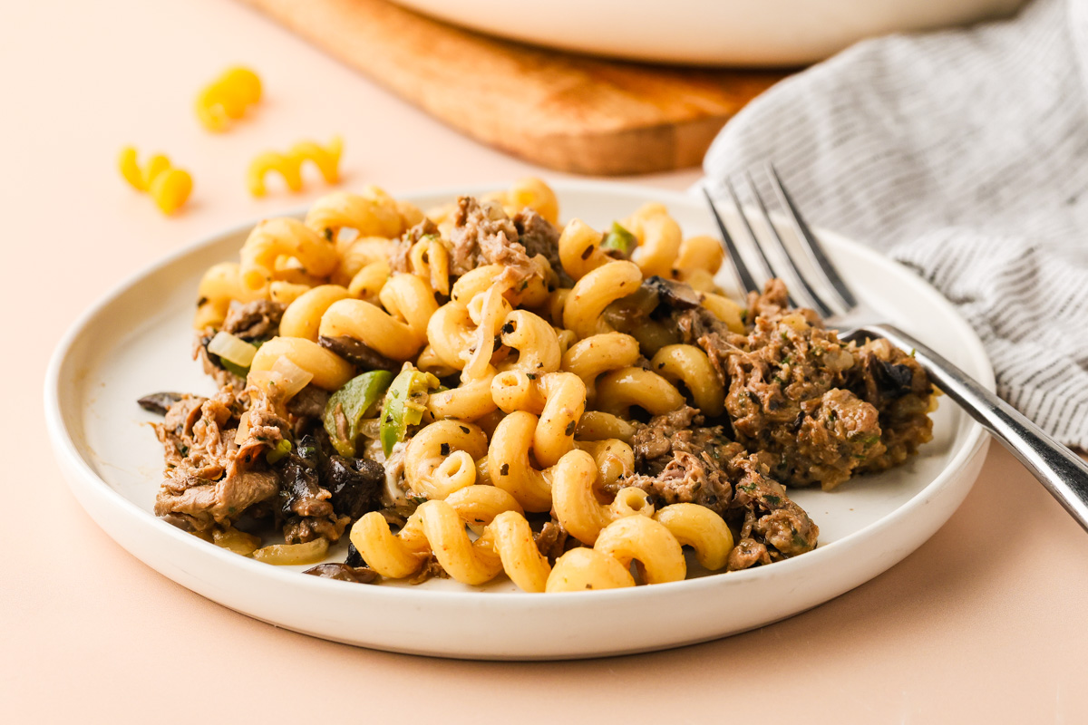 A white plate with Philly Cheesesteak Pasta- curly pasta mixed with ground beef, mushrooms, and green peppers, placed next to a fork and a striped napkin. A few uncooked pasta pieces and a wooden board are in the background.