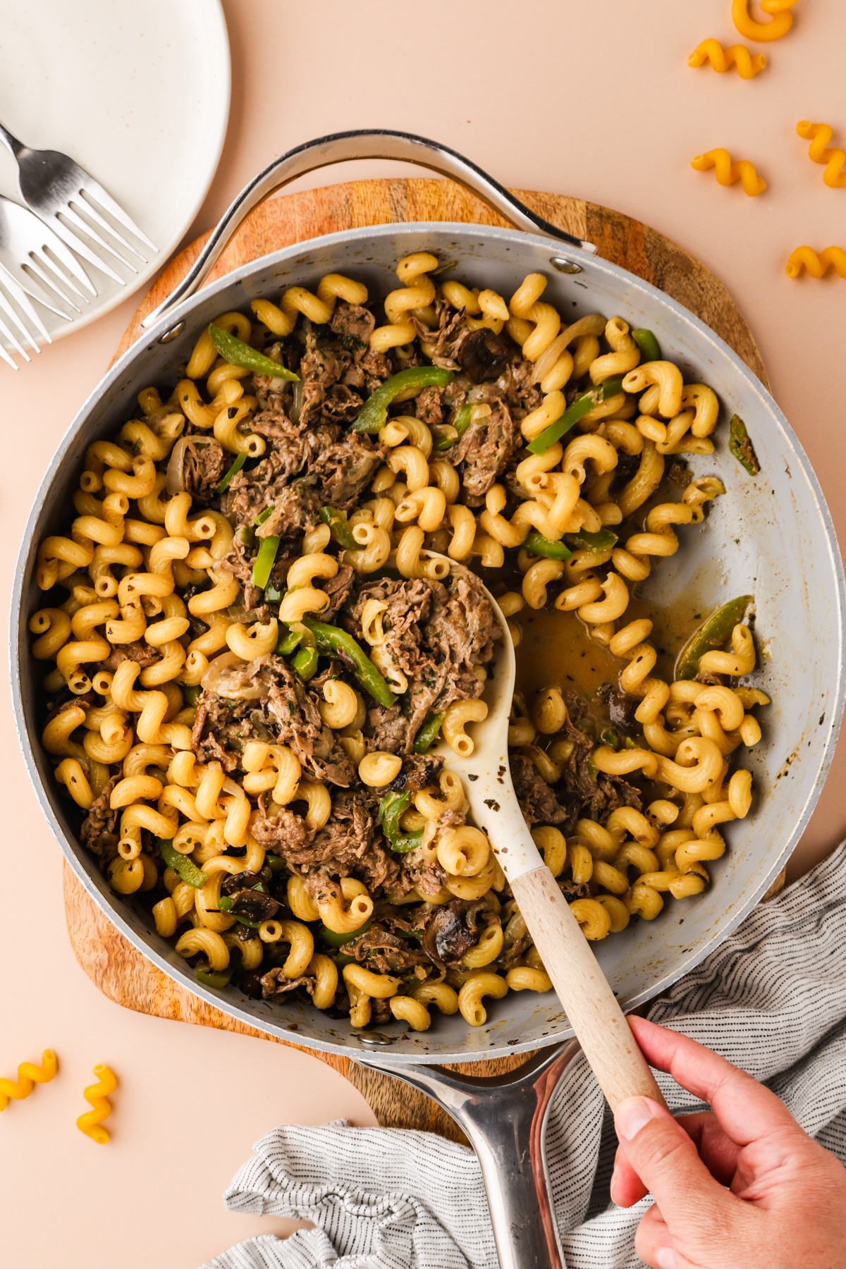 A skillet filled with Philly Cheesesteak Pasta- cooked curly pasta, shredded beef, green peppers, and mushrooms, being stirred with a wooden spoon; a plate, fork, and napkin are nearby.