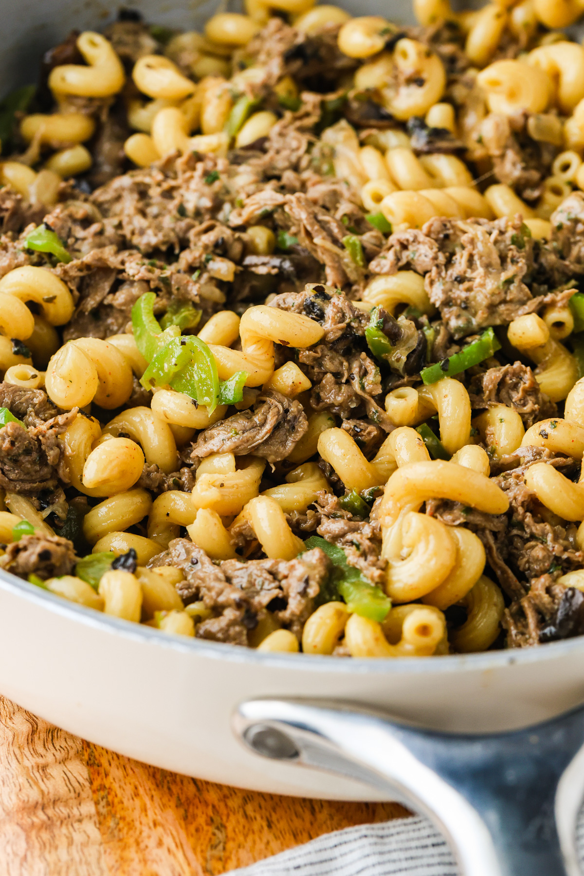 A close-up of a skillet Philly Cheesesteak Pasta- filled with cooked cavatappi pasta, pieces of beef, mushrooms, and chopped green peppers in a creamy sauce.