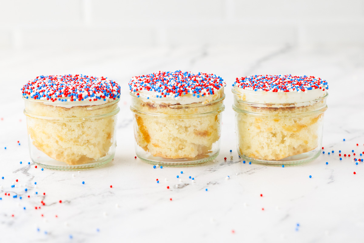 Three mini cakes in glass jars topped with white frosting and red, white, and blue sprinkles are lined up on a marble surface, with loose sprinkles scattered around.
