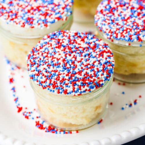 Small glass jars of cake topped with white frosting and red, white, and blue sprinkles sit on a white plate.