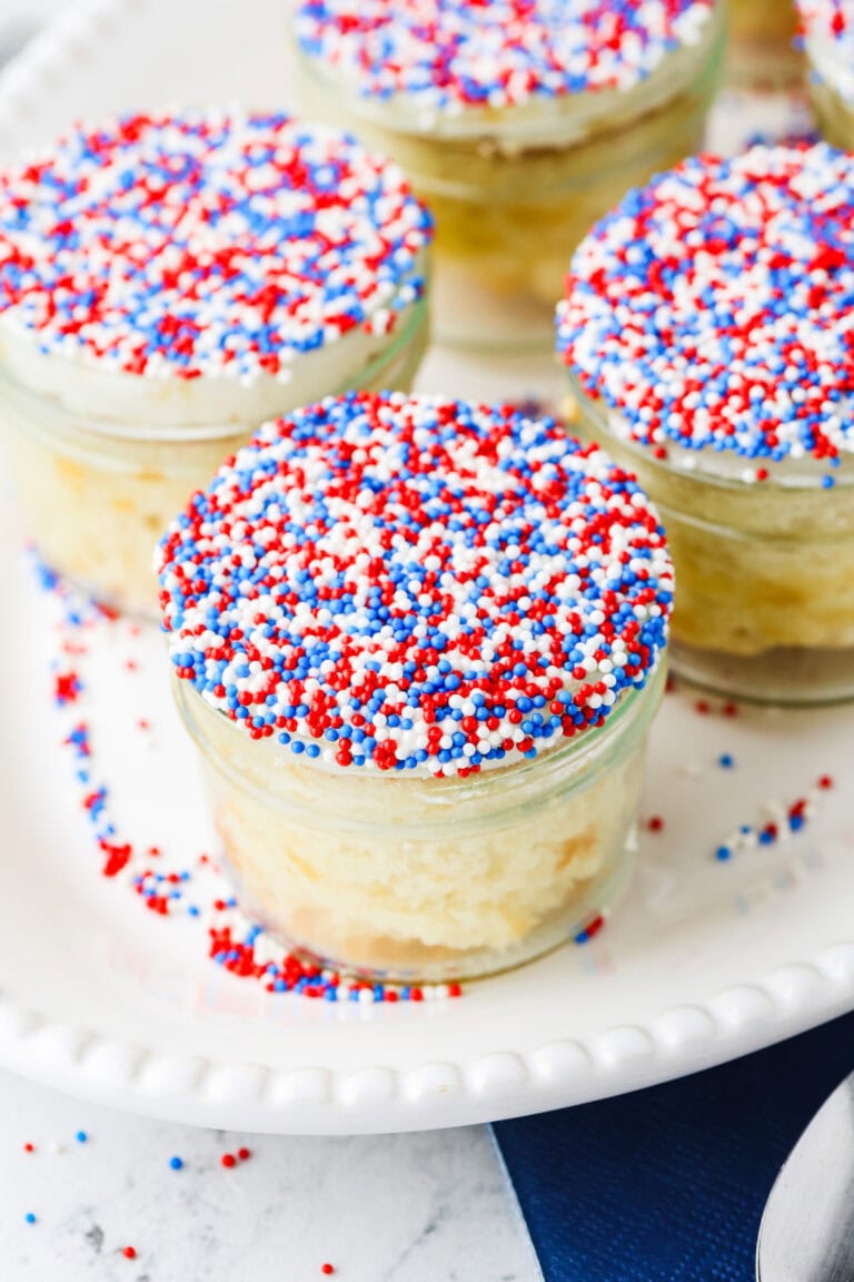 Small glass jars of cake topped with white frosting and red, white, and blue sprinkles sit on a white plate.