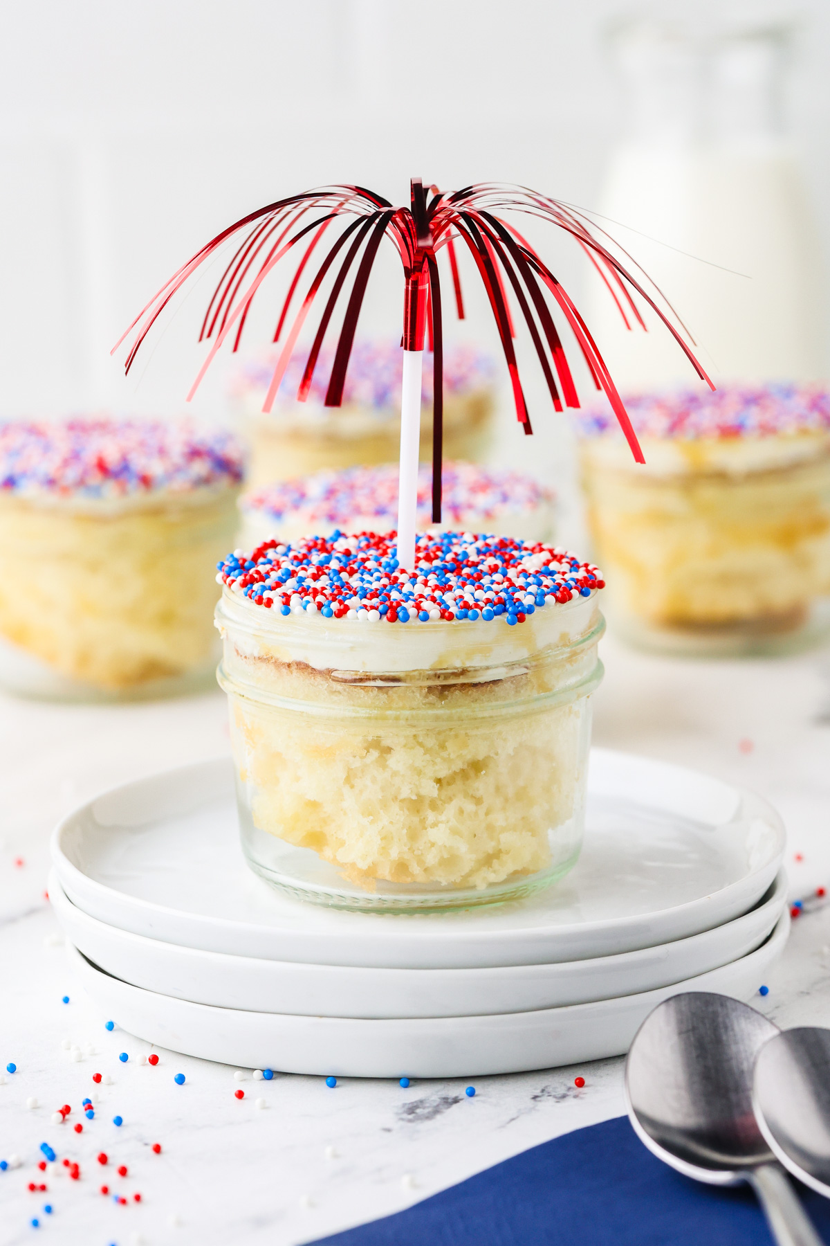 A vanilla cupcake in a jar with white frosting, red, white, and blue sprinkles, and a red metallic topper, sits on stacked white plates; other cupcakes and a milk bottle are in the background.