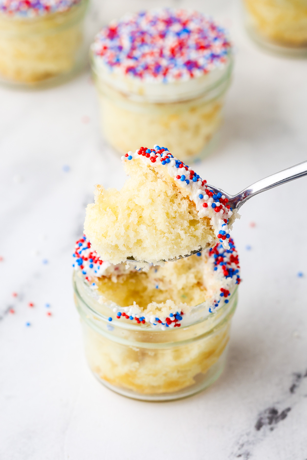 A spoon lifts a bite of vanilla cake with white frosting and red, white, and blue sprinkles from a small glass jar. More jars of similar cakes are blurred in the background.